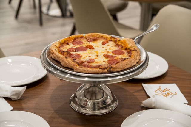 Freshly baked pepperoni pizza placed on a silver pizza stand on a restaurant table. Several white plates and folded napkins add to the dining setup, making it ideal for images focusing on food, dining experiences, or restaurant advertising.
