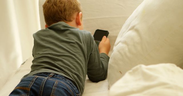 Young child lying on bed using a smartphone, dressed in a green shirt and jeans. Perfect for themes involving technology use among children, screen time discussions, home lifestyle, modern parenting, or advertising cozy home environments.