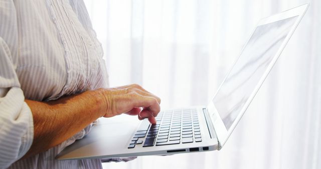 Shows senior person typing on laptop in bright natural light, suitable for illustrating themes of technology use among the elderly, modern lifestyles of older adults, internet usage, learning and working remotely.