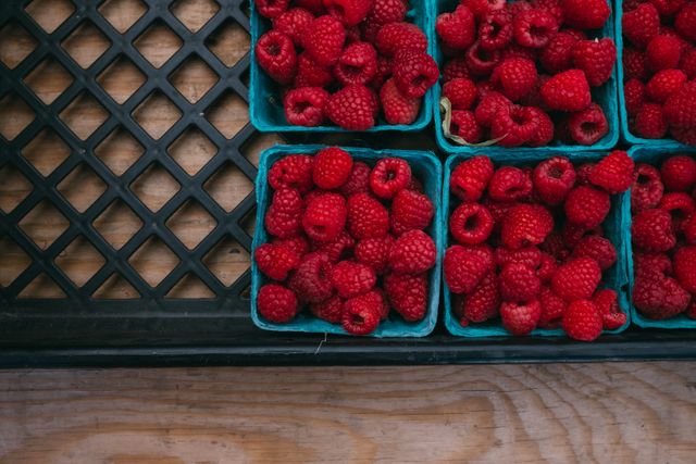 Fresh red raspberries in blue containers arranged at a market stand. Boxes are filled with vibrant red, juicy raspberries, set against a wooden and lattice background. Great for use in blogs about healthy eating, organic farming, summer recipes, and market culture. Perfect for advertisements and posters promoting fresh produce, farmers markets, or health foods.