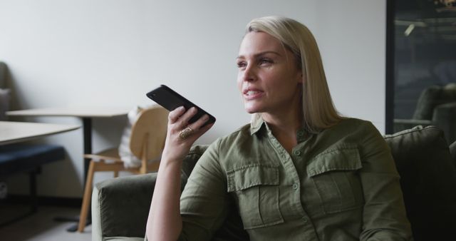 Blonde woman in a green shirt using voice command on her mobile phone while sitting in a modern office lounge. Ideal for concepts related to technology, mobile communication, modern work environments, and business technology use. Perfect for illustrating articles or advertisements focusing on tech-savvy professionals, the integration of voice command in daily work, and office lifestyle.