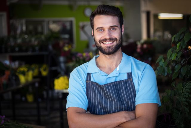 Male florist standing in a flower shop, smiling confidently with arms crossed. Ideal for use in articles or advertisements about small businesses, entrepreneurship, retail, customer service, and floral arrangements. Can also be used for promoting flower shops or illustrating professional and cheerful business owners.