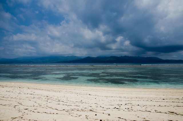 The scene shows a tropical beach with white sand and calm, clear waters under a cloudy sky. The horizon reveals distant mountains, creating a feeling of tranquility and natural beauty. This type of image is perfect for use in travel brochures, vacation advertisements, and website backgrounds related to tourism, coastal resorts, or relaxation.