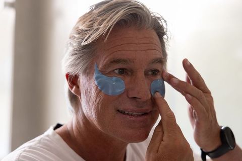 Middle-aged man applying blue hydrogel under-eye patches in bathroom mirror, skincare