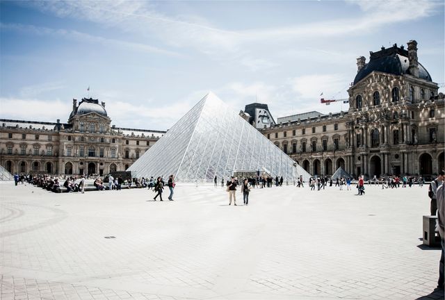 Tourists are seen exploring and walking around the courtyard of the Louvre Museum in Paris, with the iconic glass Pyramid in the center. This stock photo is ideal for use in travel blogs, tourism advertisements, educational content about French history and culture, and promotional materials for travel agencies highlighting important landmarks and tourist activities in Paris.