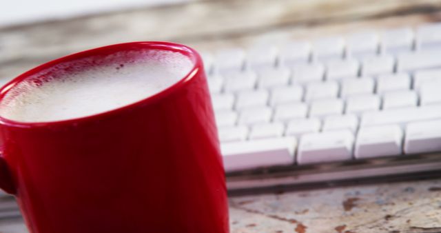 A red mug filled with a frothy beverage sits in front of a white keyboard on a desk, with copy space. It suggests a work or study environment where someone might enjoy a coffee or latte while using the computer.