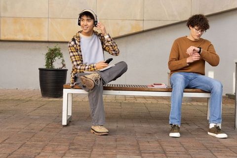 Two male students sitting on bench listening to music and checking smartwatch on campus