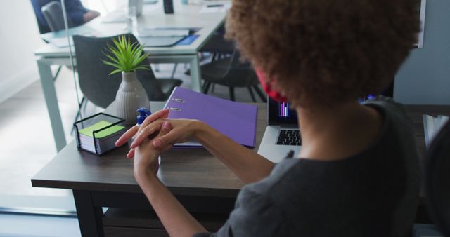 Businesswoman sitting at desk in modern office working on laptop with focused expression. Hands are clasped, suggesting deep thought or consideration. Binders, green plant, and organizational accessories on desk add to professional feel. Perfect for business, productivity, and modern office-related content.