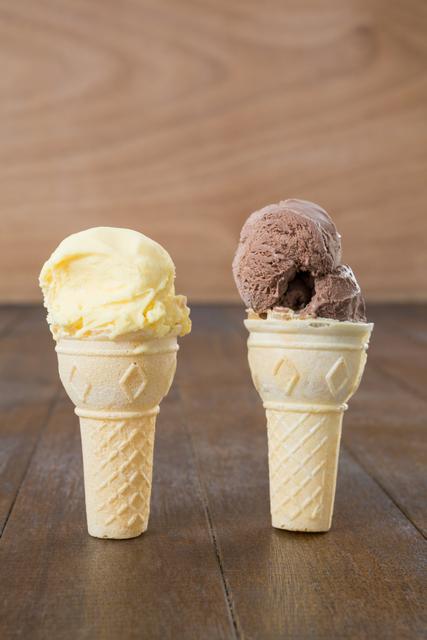 Chocolate and vanilla ice cream cones standing on a wooden surface. Perfect for use in advertisements for ice cream shops, summer promotions, dessert menus, or food blogs focusing on sweet treats and indulgent desserts.