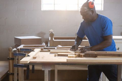 Carpenter shaping wooden planks on workbench with clamps and earmuffs in sunlit shop