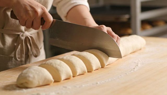 Baker slicing dough log into uniform rolls on floured wooden workbench