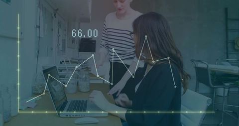 Two women collaborating over laptop with line graph overlay in modern office