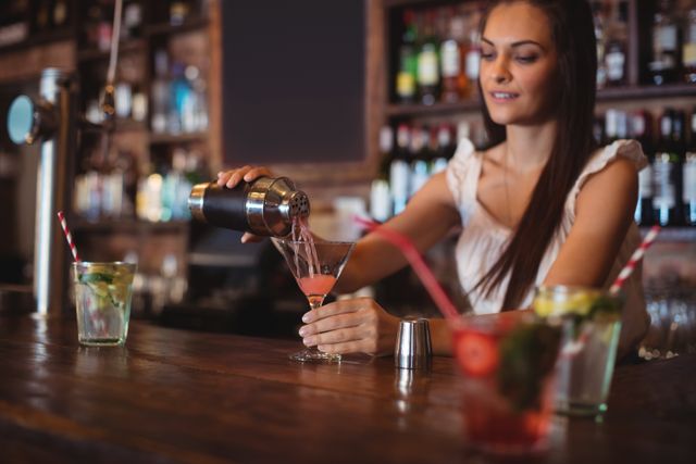 Female bartender pouring a cocktail into a glass at a bar counter. Ideal for use in articles or advertisements related to nightlife, bar services, mixology, and hospitality industry. Can also be used for promoting bars, restaurants, and cocktail recipes.