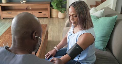 African american caregiver checking blood pressure of mature woman on living room sofa