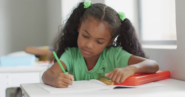 Young African American girl writing at a desk in a classroom setting, holding a pen and concentrating on her work. Ideal for educational websites, school promotions, materials on childhood education, and academic advertisements.