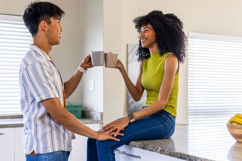 Smiling multicultural couple toasting coffee while sitting on kitchen counter