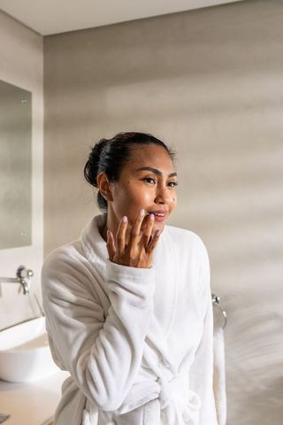 Asian woman applying skincare in modern bathroom wearing white bathrobe, serene self-care