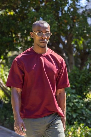 African american man walking in sunlit garden wearing maroon tee, sunglasses and smoking