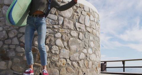 Woman balancing on skateboard holding surfboard at seaside promenade
