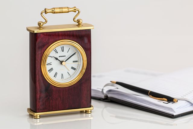 Ideal for illustrating themes of time management, productivity, and office aesthetics. This vintage desk clock with a wooden frame and golden detailing complements the open planner and pen, making it perfect for business or organizational-related content. Suitable for websites, blogs, or advertisements focusing on workplace environments, efficiency tips, or time tracking tools.