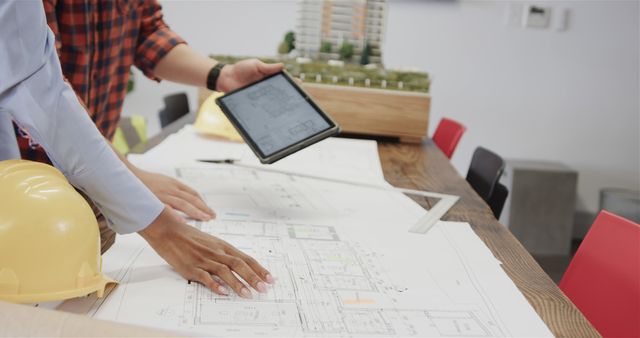 Architects are discussing building plans, using a digital tablet and blueprints. A yellow hard hat rests on the table, suggesting safety measures on a construction project. An architectural model in the background adds to the focus on design and planning. This can be used for articles or content about architecture, engineering, construction projects, professional collaboration, and team planning.