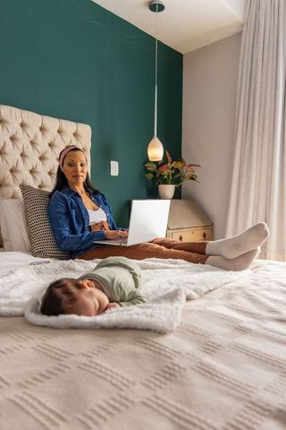 African american mother working on laptop while baby sleeping on bed in cozy green bedroom