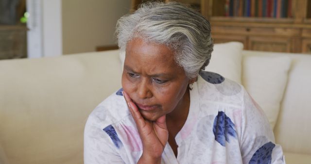 Senior woman with grey hair sitting on couch in living room, looking pensive and thoughtful. Great for use in articles about aging, mental health, solitude, elderly care, and reflective moments in senior citizens' lives.