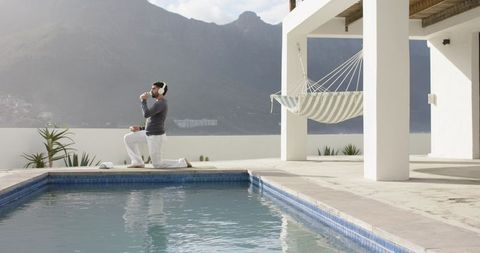 Indian man kneeling and sipping water by pool wearing headphones with hammock and mountain coastline