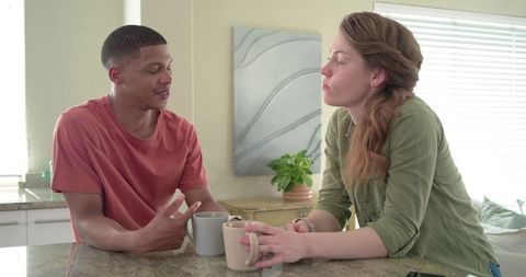 Diverse couple leaning on kitchen island holding mugs and chatting over morning coffee