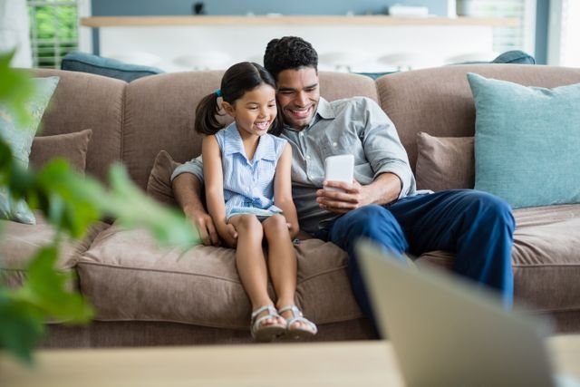 Father and daughter sitting on sofa, smiling while looking at mobile phone. Perfect for illustrating family bonding, parenting, and technology use in everyday life. Ideal for advertisements, blogs, and articles related to family life, parenting tips, and home technology.