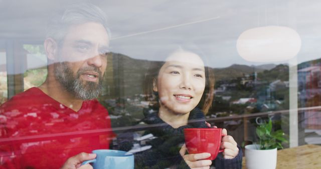 Couple standing inside home, holding coffee cups, smiling while looking out window at scenic view. Useful for promoting relationship goals, cozy morning routines, home decor, and lifestyle content emphasizing happiness and connection.