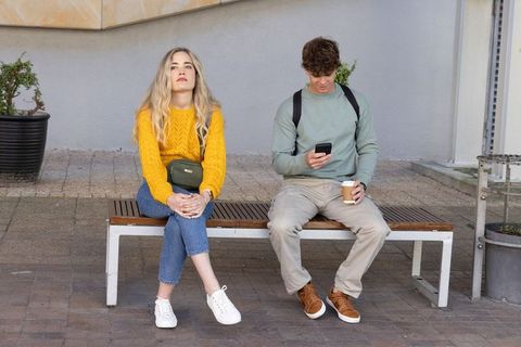 Young adults sitting on bench using smartphone and drinking coffee, urban contrast