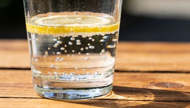 Close-up sparkling water with lemon slice in glass on wooden table, sunlit refreshing drink