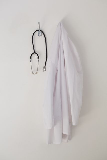 Laboratory coat and stethoscope hanging on a hook against a white wall. Ideal for use in medical, healthcare, and professional settings. Suitable for illustrating concepts related to doctors, nurses, hospitals, clinics, and medical equipment.