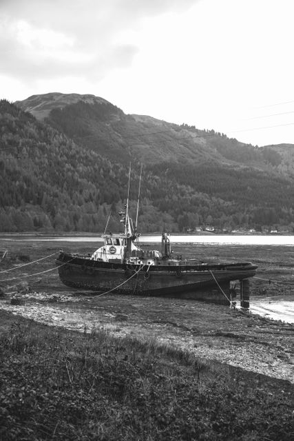 Abandoned boat resting on the shore during low tide in a coastal area surrounded by mountains. Monochrome captures the rustic and serene atmosphere. Ideal for themes on abandonment, nature's passage of time, or maritime history.