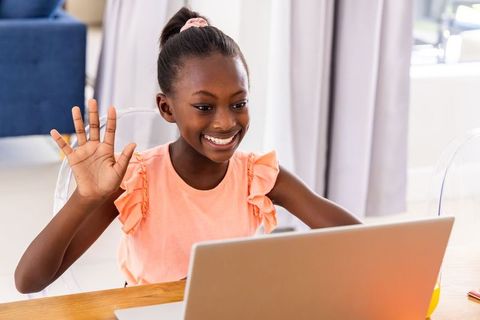 Smiling african-descent girl waving during video call on laptop in bright home interior