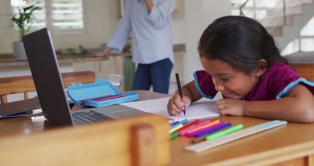 Young girl focused on doing homework at home, sitting at table with laptop, colorful pencils, and ruler. Father standing in background talking on phone. Ideal for content about home education, remote learning, child development, and family life during remote schooling.