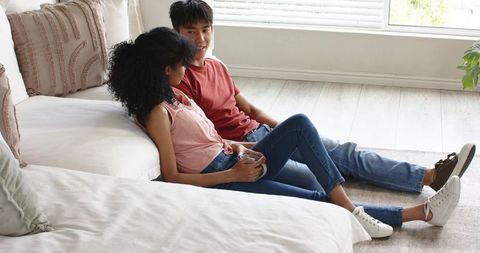 Mixed-race couple relaxing on floor leaning against sofa sharing coffee in bright minimal living roo