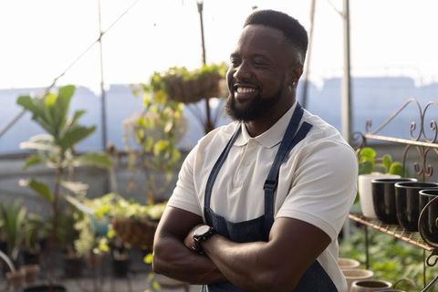 Smiling african american gardener wearing denim apron and watch in sunlit greenhouse