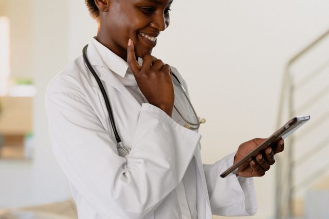 African American female doctor smiling while using a tablet. She is wearing a white coat and a stethoscope, indicating her medical profession. This image can be used for healthcare websites, medical blogs, telemedicine services, and promotional materials for hospitals and clinics.
