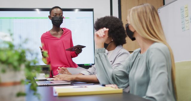 Three diverse businesswomen wearing face masks in discussion at office meeting, one holding tablet. work at an independent creative business during covid 19 coronavirus pandemic.
