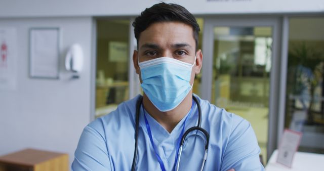 Portrait of biracial male doctor wearing scrubs and face mask standing in hospital. medicine, health and healthcare services during covid 19 coronavirus pandemic.