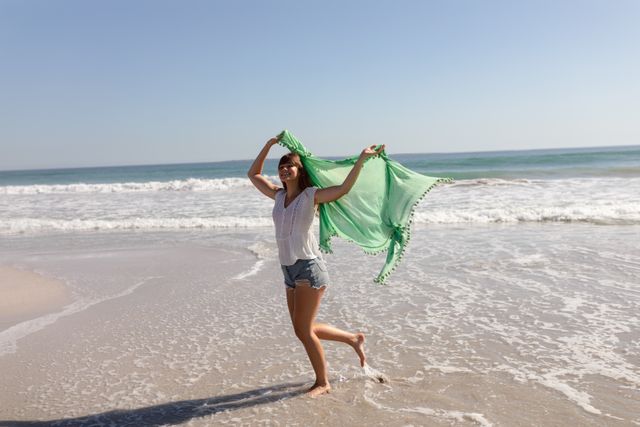 Young woman waving green scarf on beach, enjoying sunny day. Perfect for travel blogs, summer vacation promotions, lifestyle articles, and advertisements focusing on relaxation and outdoor activities.