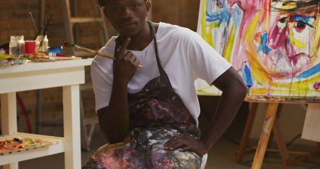 Portrait of african american male artist wearing apron holding a paint brush smiling at art studio. art, hobby and creative occupation concept