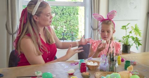 Mother teaching daughter decorating easter eggs at sunlit kitchen table for spring celebration