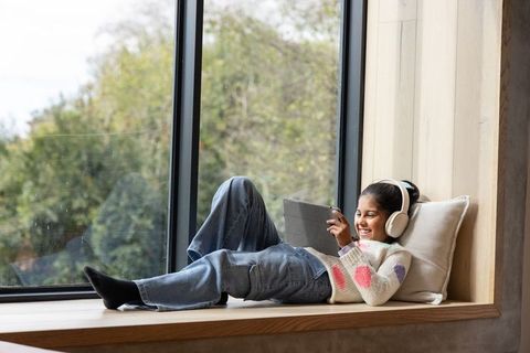 Indian preteen girl lying on window seat holding tablet wearing headphones, smiling