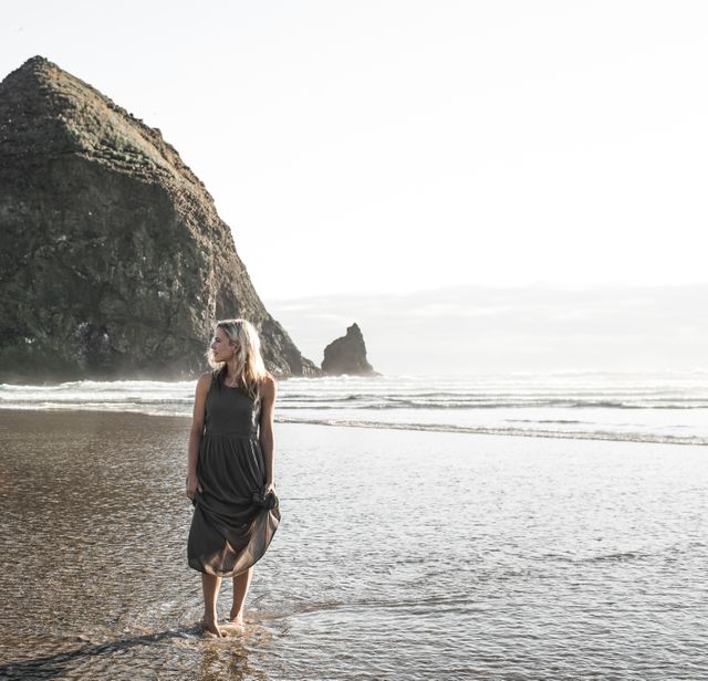 Blonde woman standing in shallow water near a rocky shoreline. Perfect for themes of nature, serenity, coastal living, summer vacations, and peaceful retreats. Useful for travel blogs, lifestyle articles, and promotional material for beach resorts.