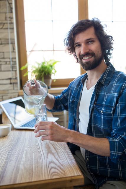 Man pouring lemonade into a glass while sitting at a wooden table with a laptop in a bright, cozy home environment. Ideal for use in lifestyle blogs, home living articles, or advertisements for beverages and home products.