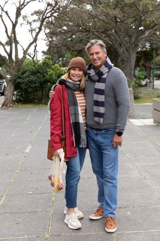 Middle-aged couple smiling on sidewalk in casual fall knitwear holding mesh bag of apples