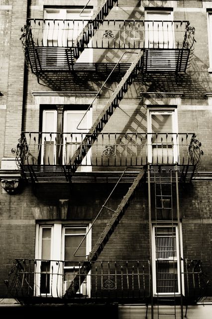 This image features a classic fire escape on the facade of a historic urban building. The black-and-white filter adds a vintage feel, highlighting the iron railing and intricate design. Ideal for use in topics related to city life, urban planning, safety, architecture history, or vintage aesthetics.